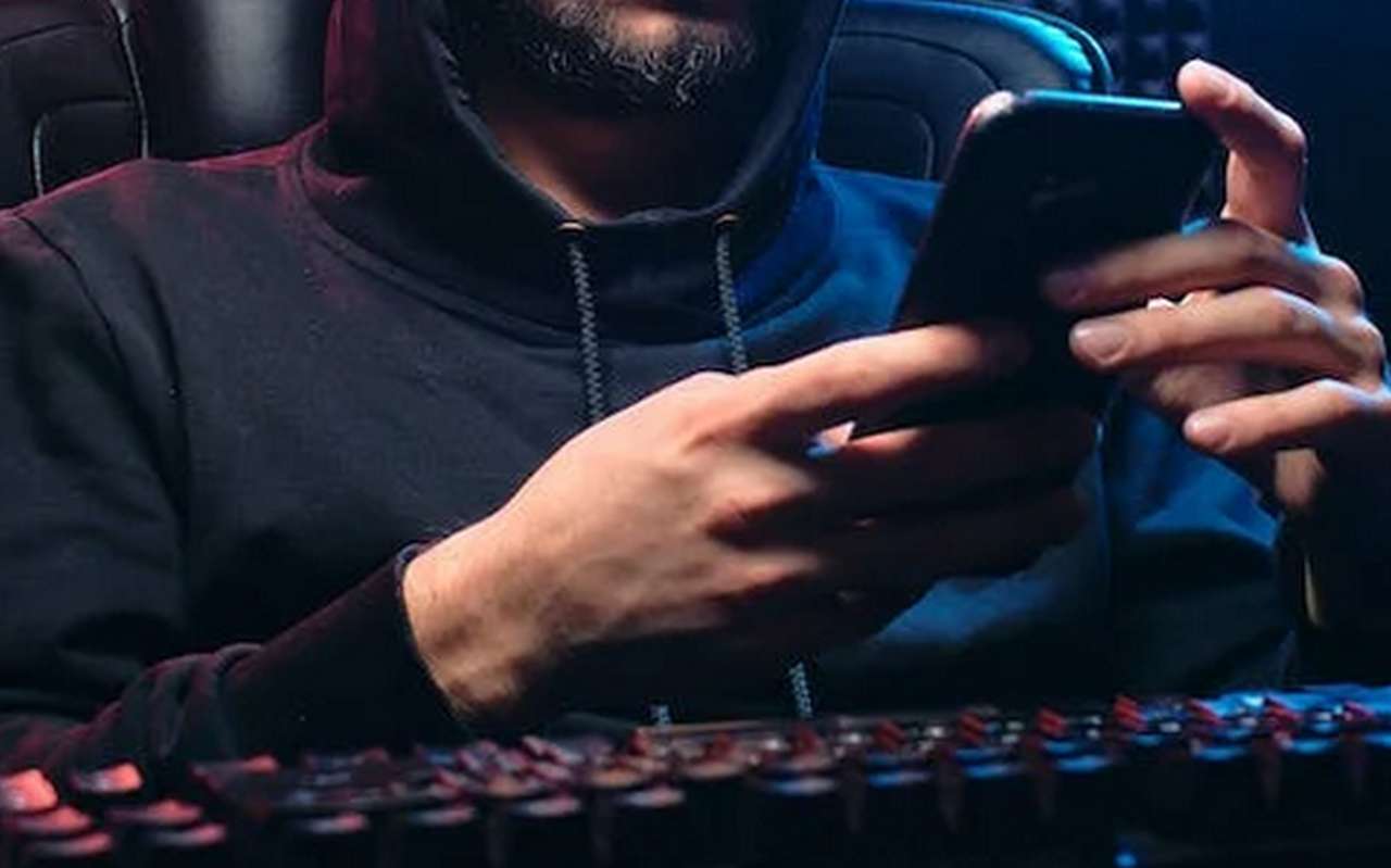 un uomo con felpa e cappuccio alla tastiera di un pc con il cellulare in mano